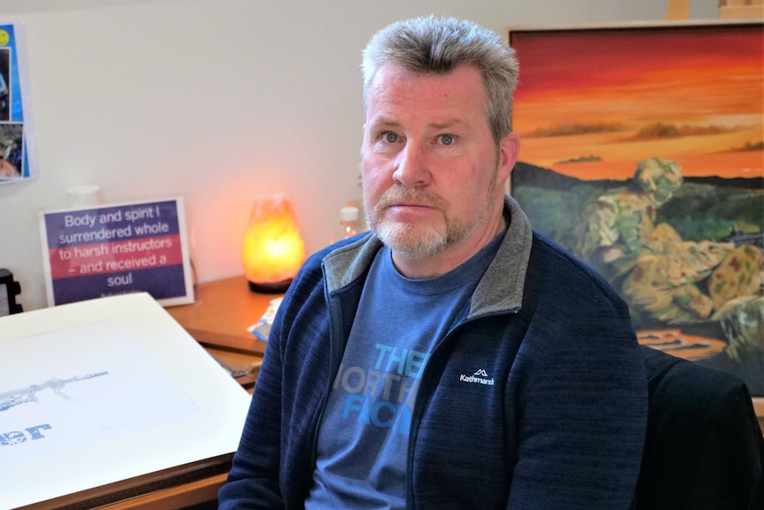 A middle aged man sits in front of a desk and military paintings.