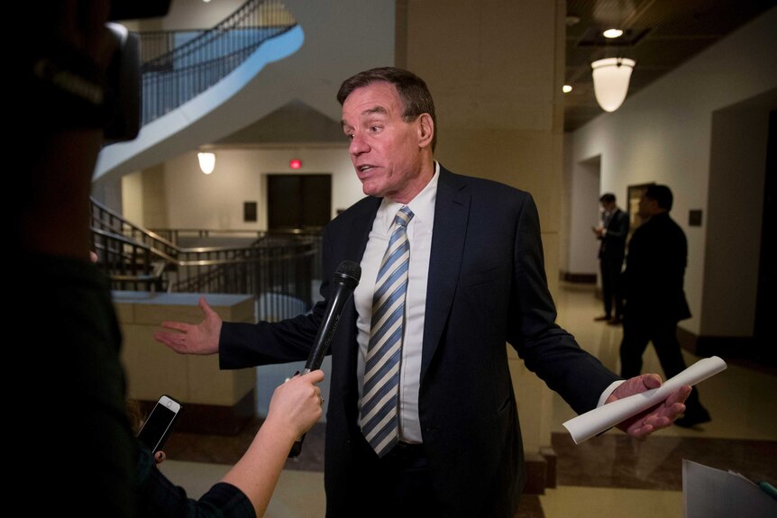 US Senator Mark Warner opens his arms as he addresses one reporter with a microphone while wearing a dark suit.