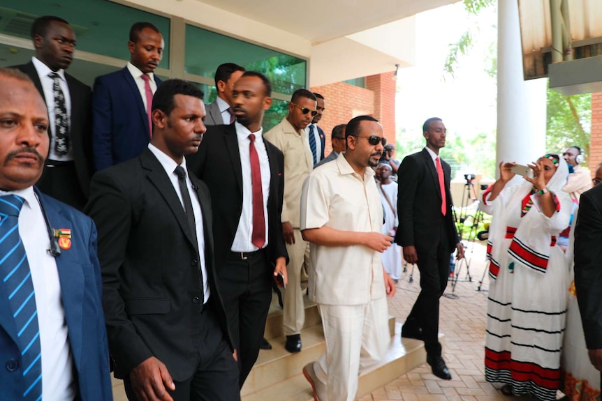 A crowd of men walk down small steps in a building corridor, with the Ethiopian PM at its centre, dressed in white.
