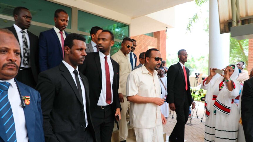 A crowd of men walk down small steps in a building corridor, with the Ethiopian PM at its centre, dressed in white.