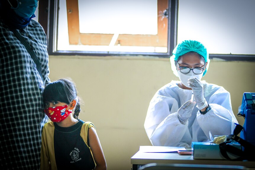 A little girl in a Hello Kitty face mask leans against a woman as a nurse in scrubs prepares a needle