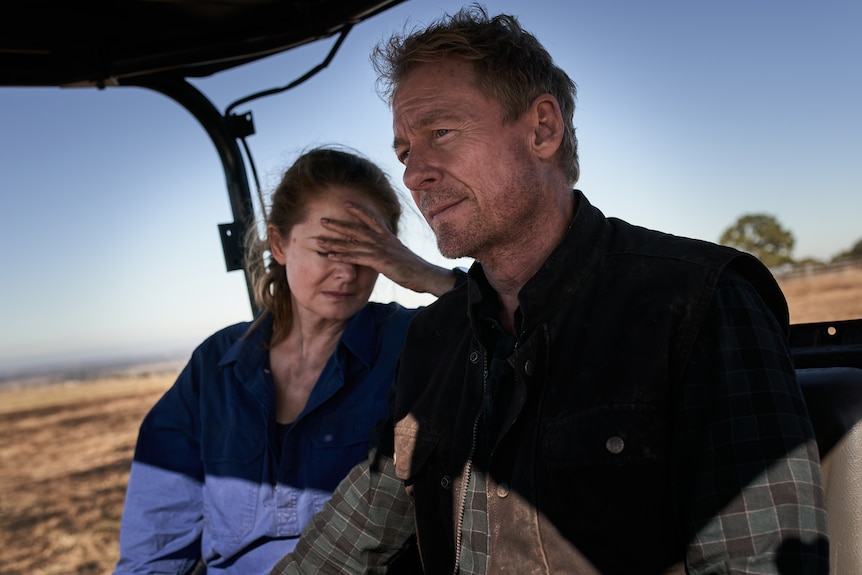 Woman with hand to head and man in cabin of tractor with rural background.