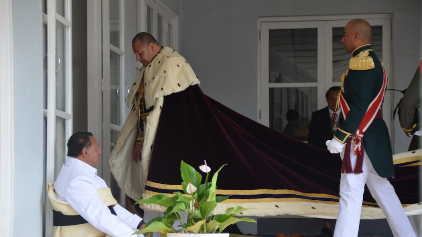 King Tupou VI wearing a cloak walks into a building while a man sits down in the foreground and an attendant walks behind.