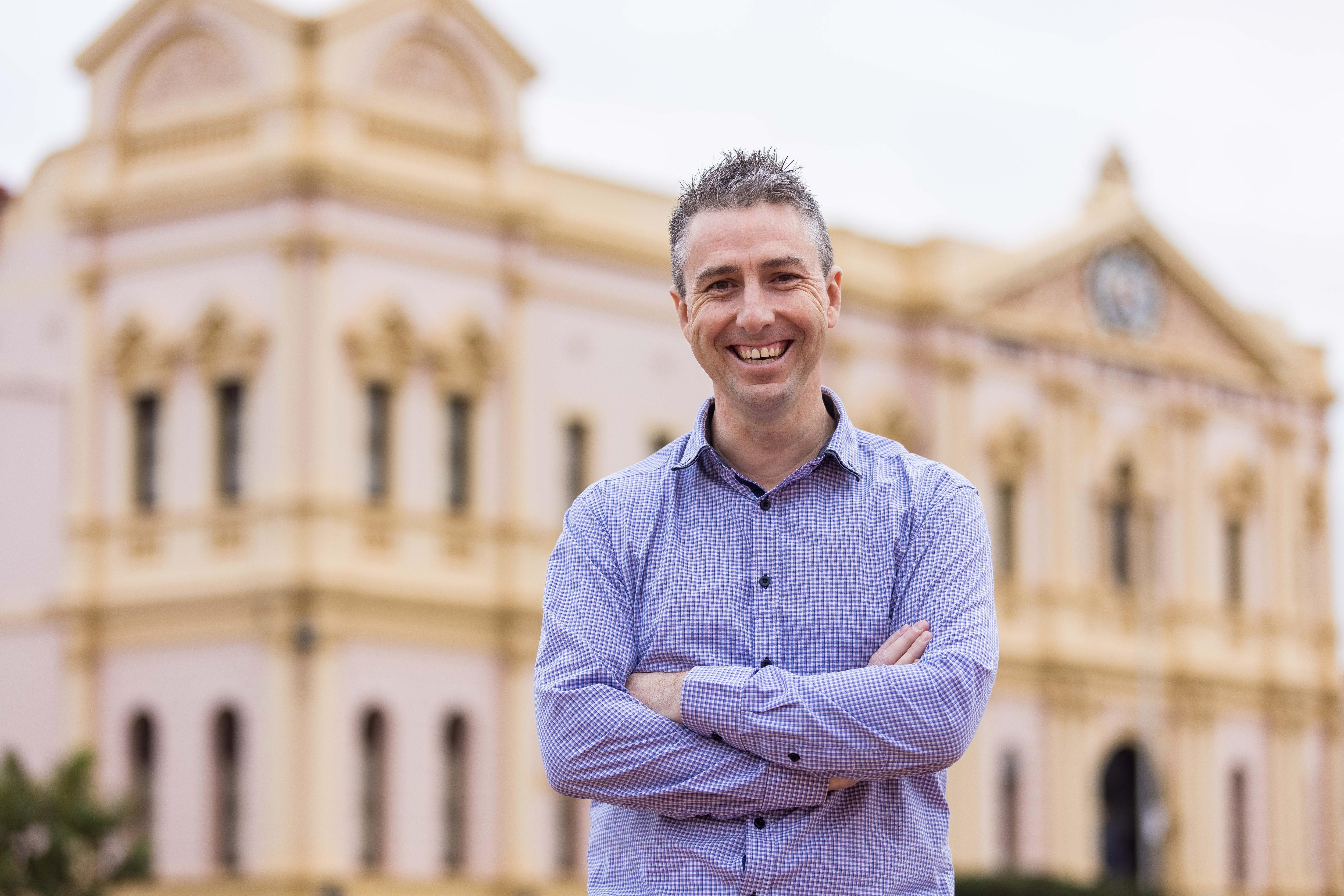 A man in a business shirt with arms crossed in front of a heritage building.  