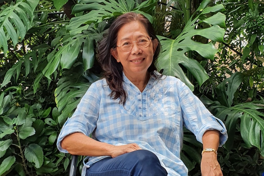 Auntie Hong sits in front of monstera plants and smiles. She has mid-length dark hair, wears round glasses and blue gingham top