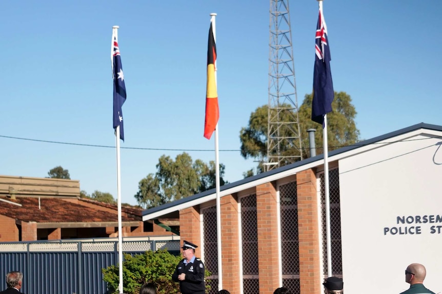 A police officer speaking to a crowd of people in front of three flags outside Norseman police station.