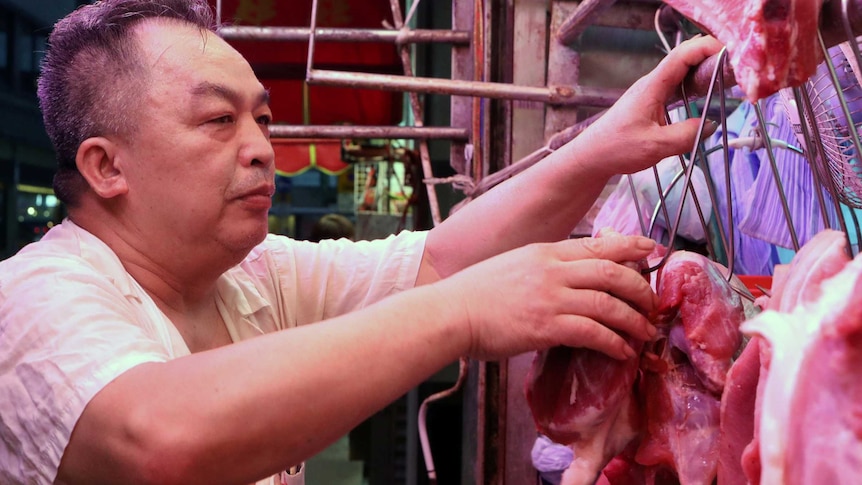 A vendor touches a piece of meat hanging from a hook at a market