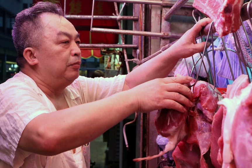 A vendor touches a piece of meat hanging from a hook at a market