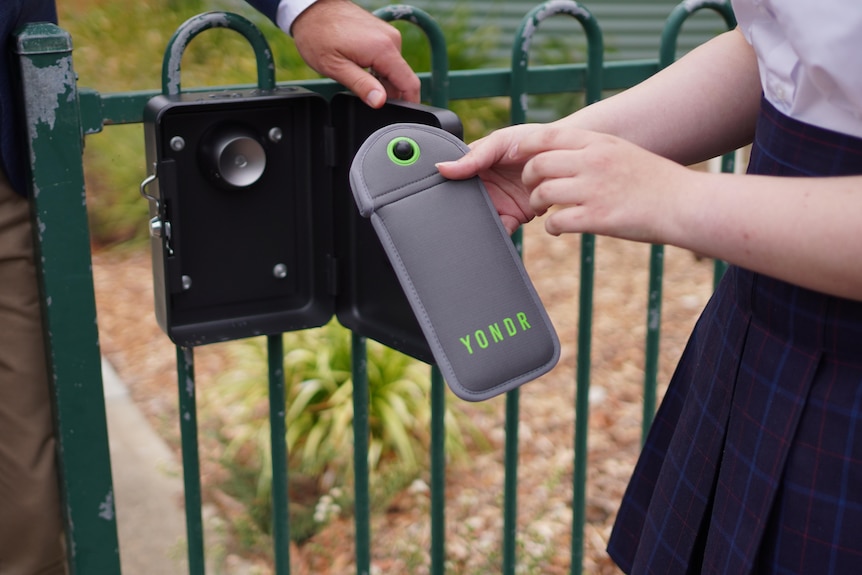 A close-up of a student's hands tapping a grey pouch against a black box on a green school fence