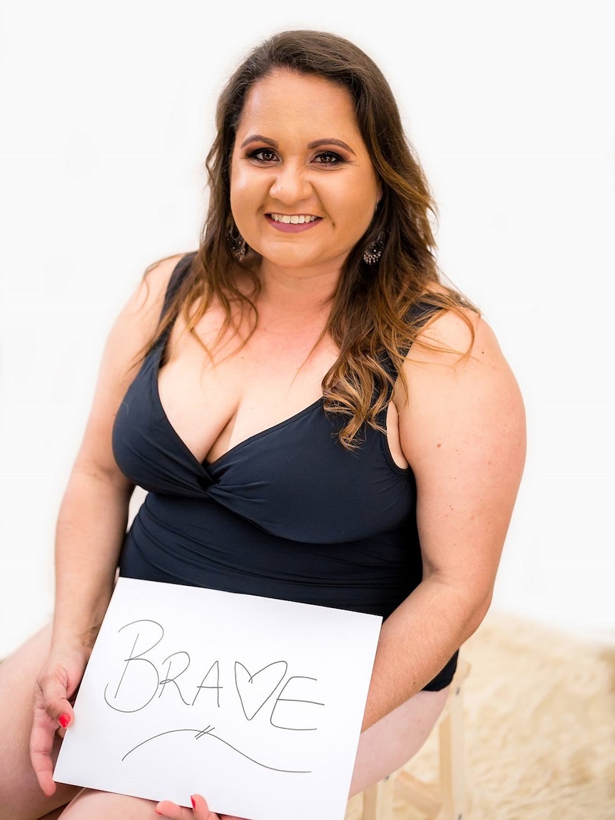 A woman in a singlet and underwear sits on a chair holding a brave sign.