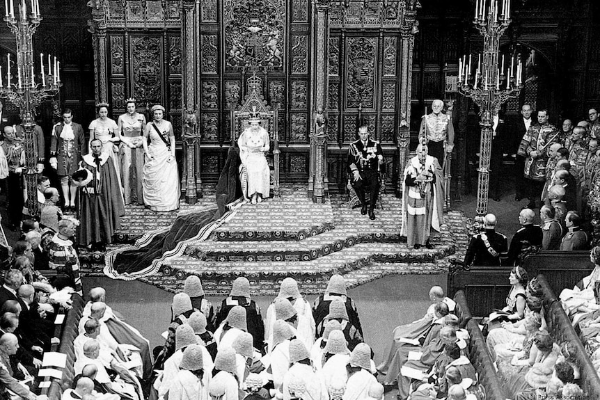 A black and white photo of the Queen sitting on a throne in the centre of a large room surrounded by people in formal uniforms.