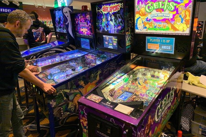 Looking over a man's shoulder as he concentrates on playing a pinball machine.