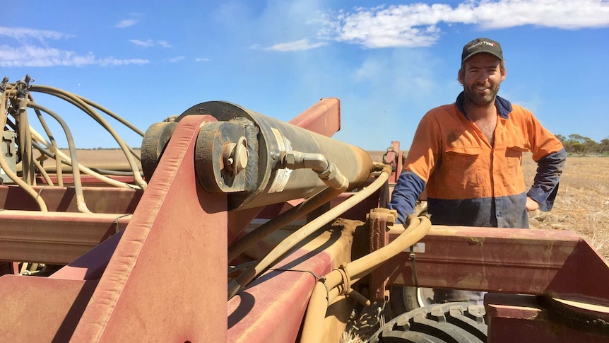 A man wearing a hat and high-viz shirt smiles as he stands next to farm machinery in a field.