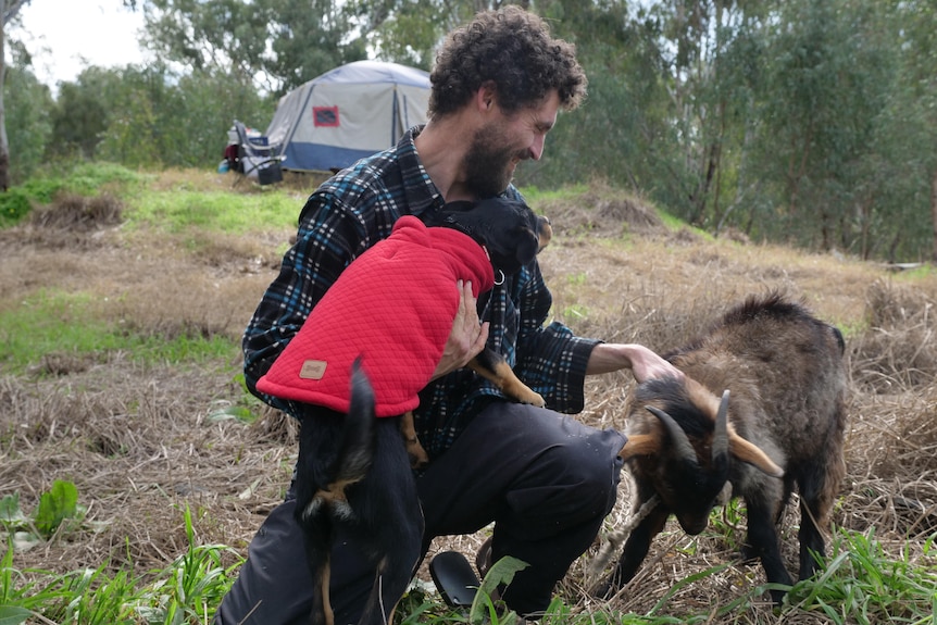 A man smiles at his goat and dog in front of a tent on a hill.