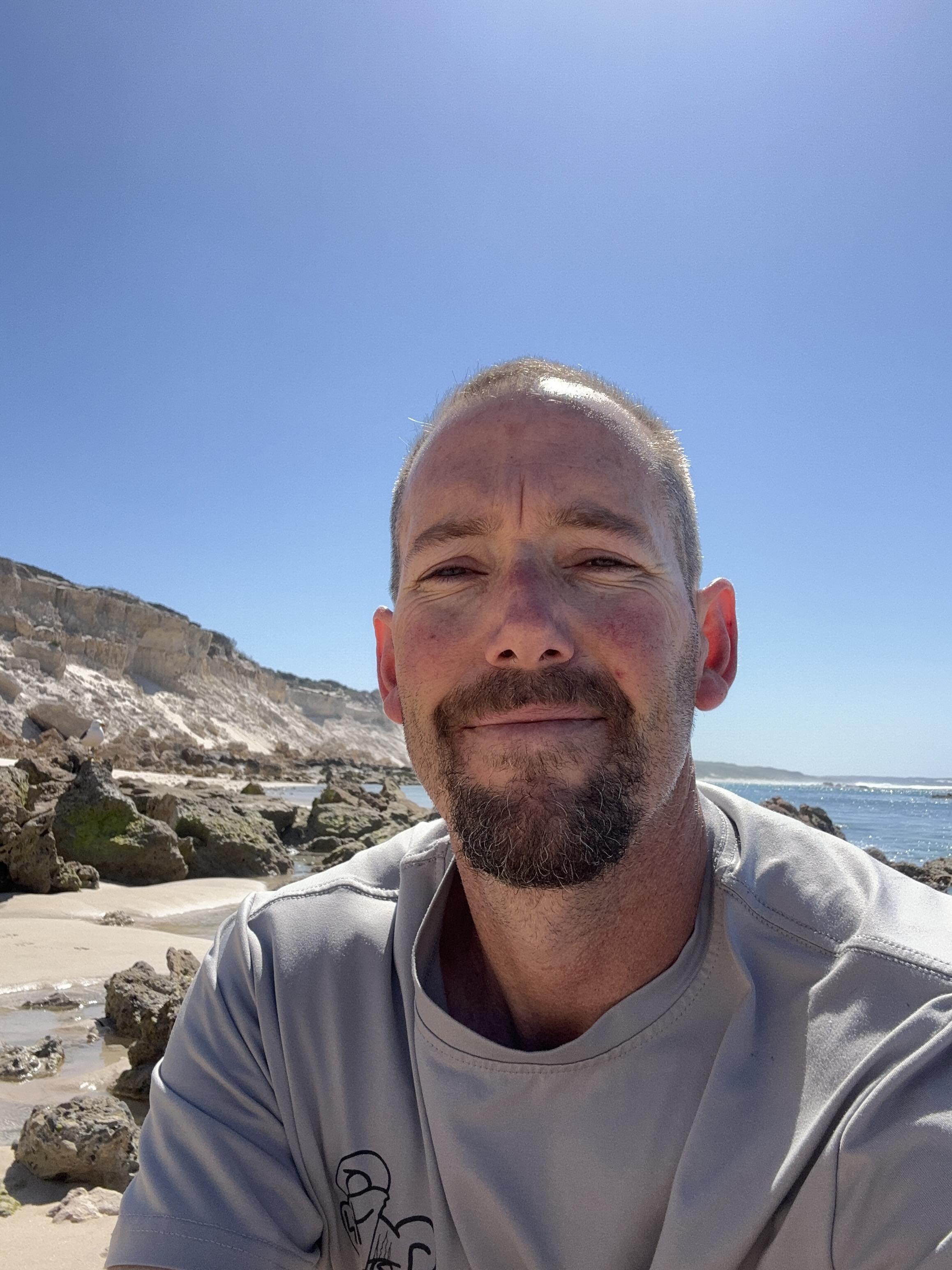 A headshot of man. Hee stands in the sun, has a moustache and goatee and wears a t-shirt.