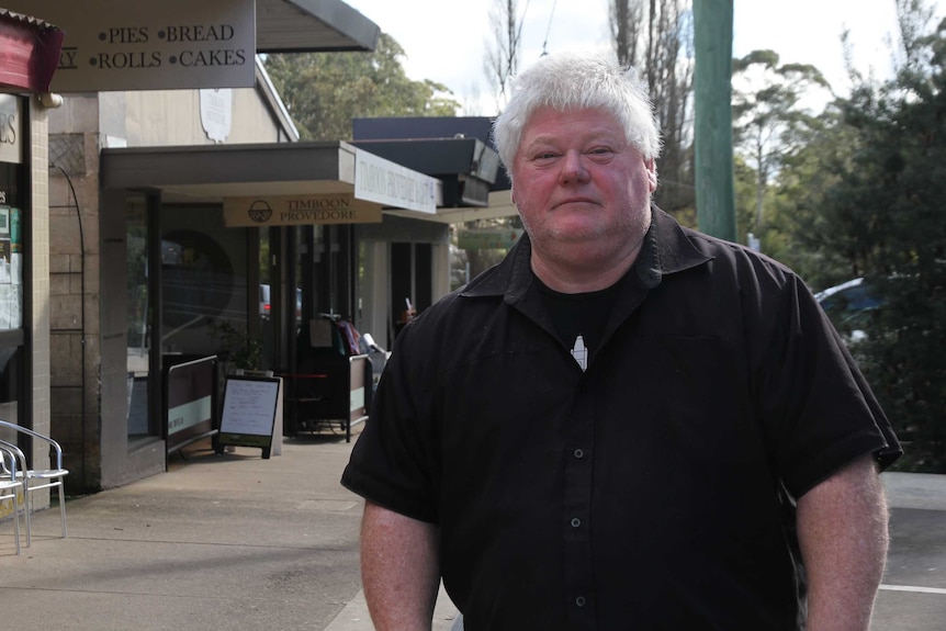 A stout man in his 50s stands outside shops in a country town.