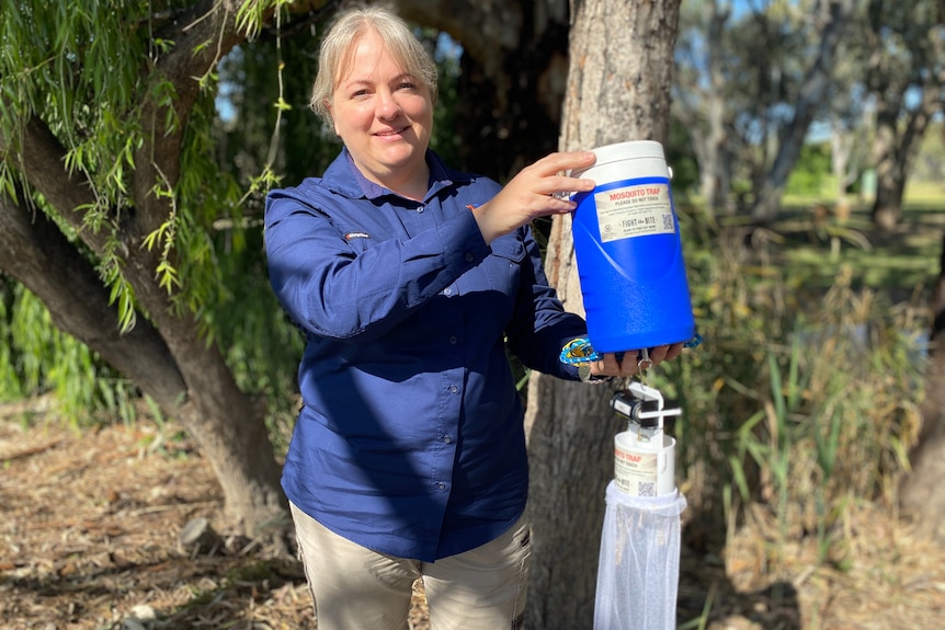 A woman with blonde hair smiles, holding a blue esky mosquito trap, standing in trees