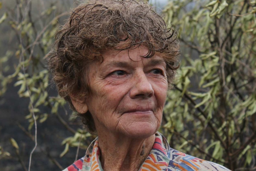 A older woman with curly hair stands in front of black charred landscape