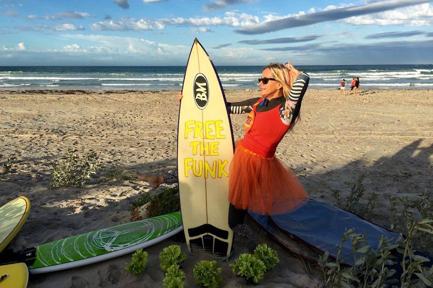 Rose Holdaway, wearing a red tutu, on a beach with a surfboard with the words "Free the Funk" written on it.