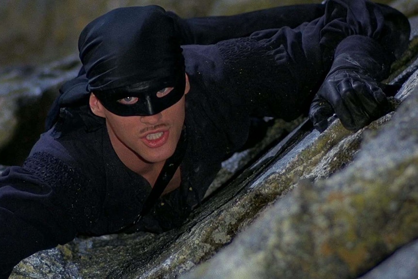 A man wearing black clothes, black gloves, a black bandana and a black domino mask scales a cliff.