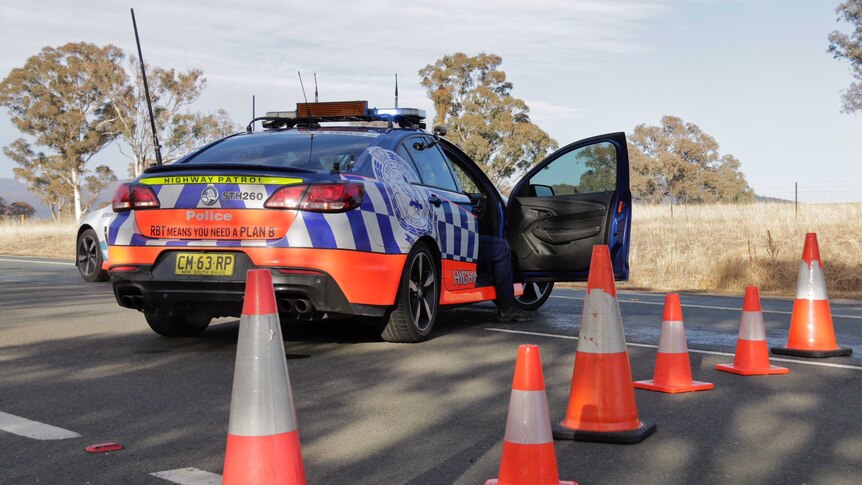 A police officer's leg can be seen out of the open door of a police car sitting behind traffic cones on a highway.