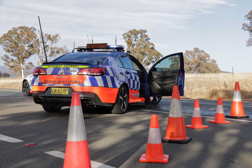 A police officer's leg can be seen out of the open door of a police car sitting behind traffic cones on a highway.