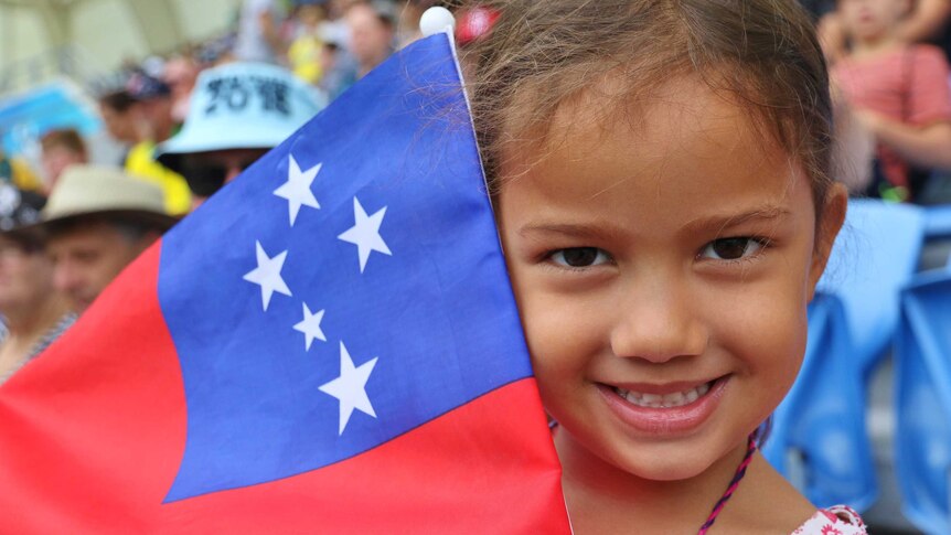 A little girl smiles at camera with a Samoan flag held beside her face.
