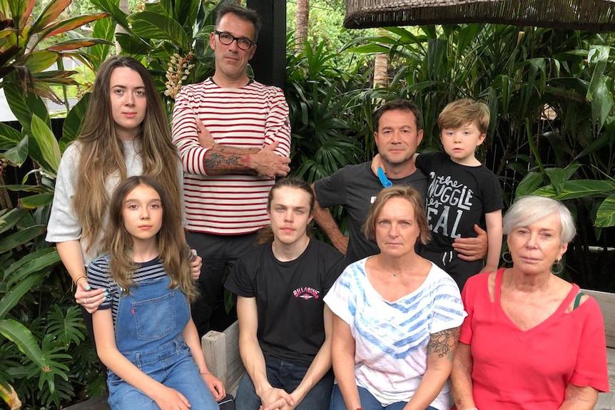 A group family photo with 8 people outside. Greenery behind.