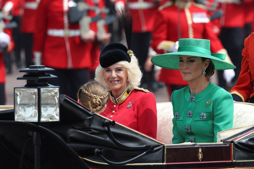 A woman in a green dress and hat sits next to a woman in a red coat and hat.