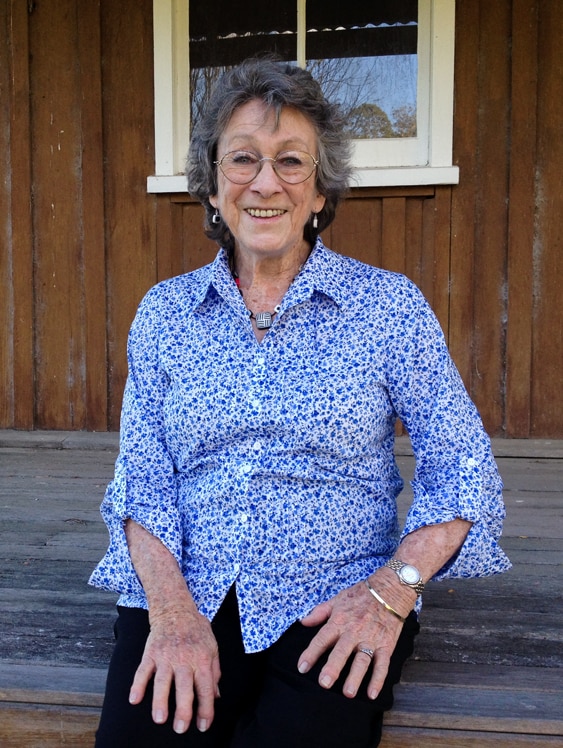 A smiling, older woman – Joy McKean – sits on the veranda at a country homestead.