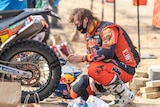 A man in orange motorcycle clothing squats next to a motorcycle.