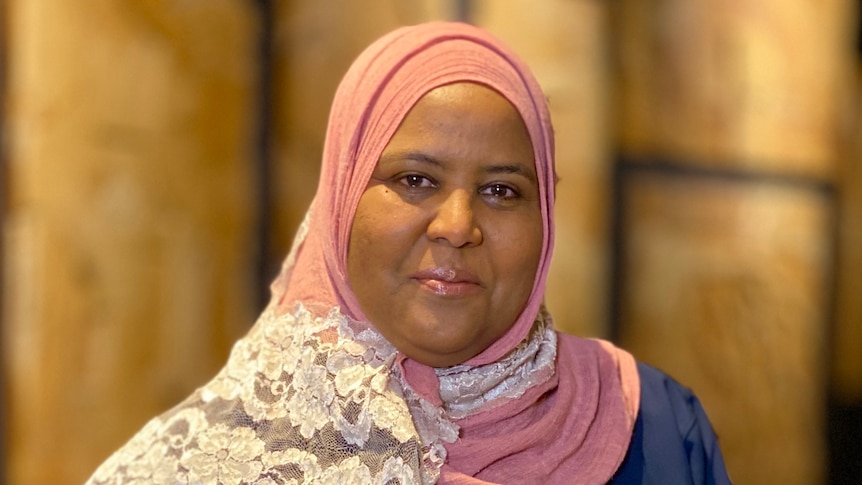 Nagat Abdalla proudly holds a cookbook, dressed in a pale pink hijab with a latticed design.