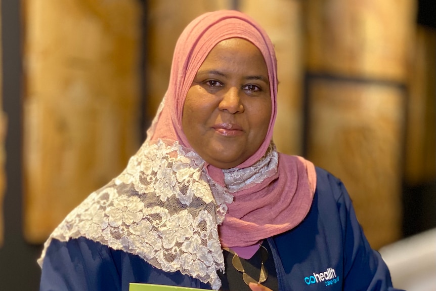 Nagat Abdalla proudly holds a cookbook, dressed in a pale pink hijab with a latticed design.