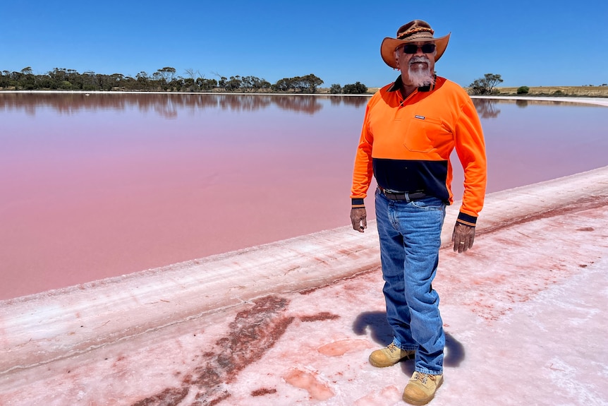 Aboriginal elder standing in front of pink lake with blue sky wearing jeans, orange shirt, hat and sunglasses 