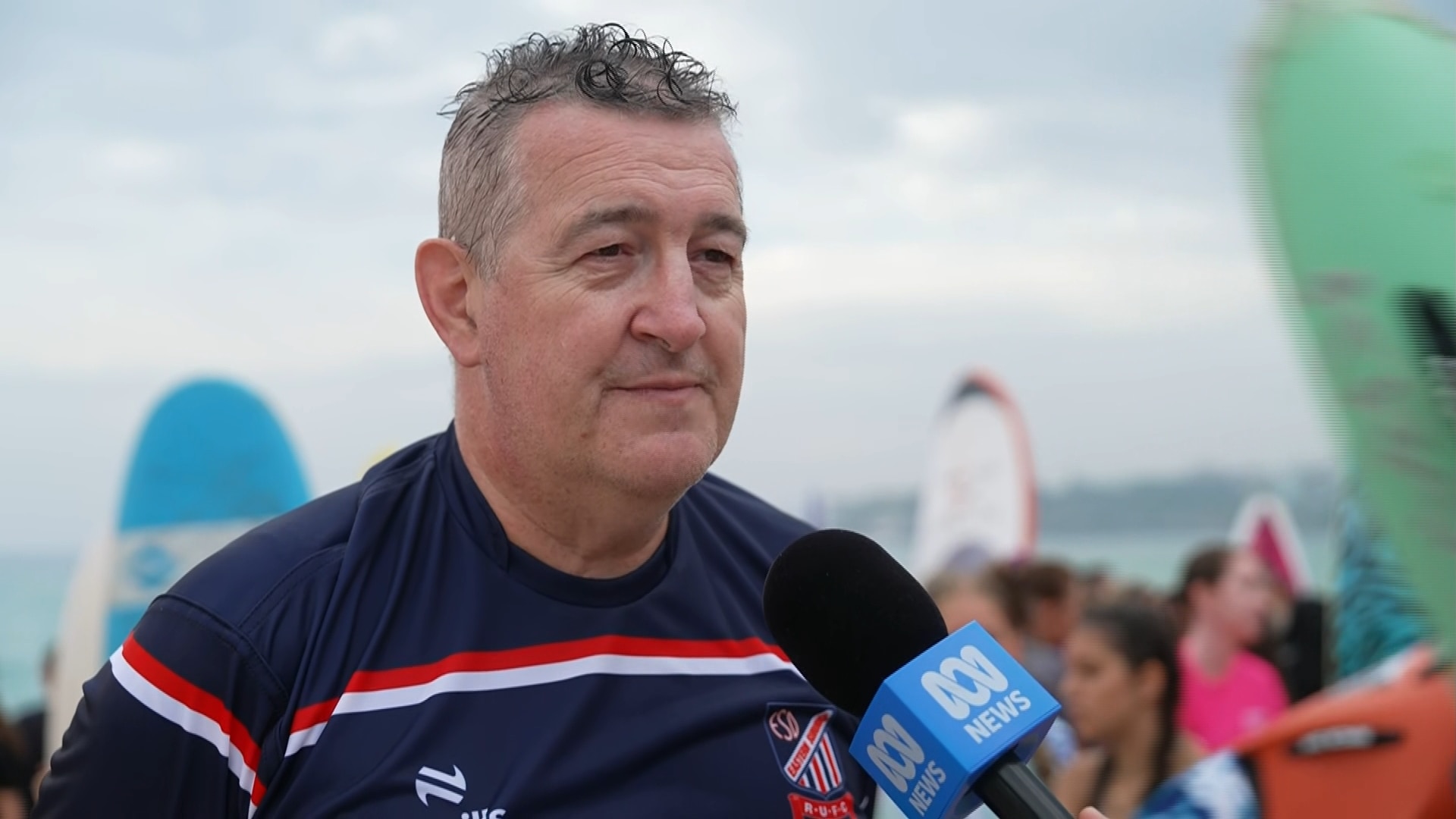 O Sr. Jackson está vestindo uma camisa de rugby com pranchas de surf e pessoas ao seu redor na praia.