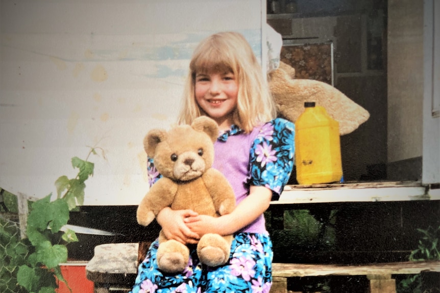 A girl with blonde hair holds a bear and smiles