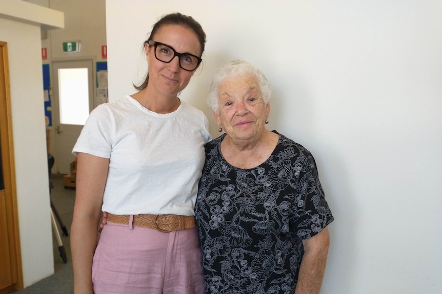 One older woman and younger women stand next to each other against a wall. Older woman has grey hair, wears black printed dress.