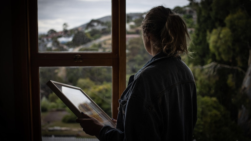 An unidentified woman holds a picture frame and looks out a window