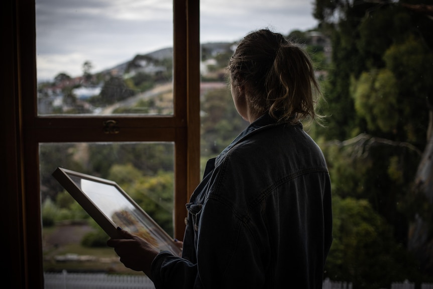 An unidentified woman holds a picture frame and looks out a window