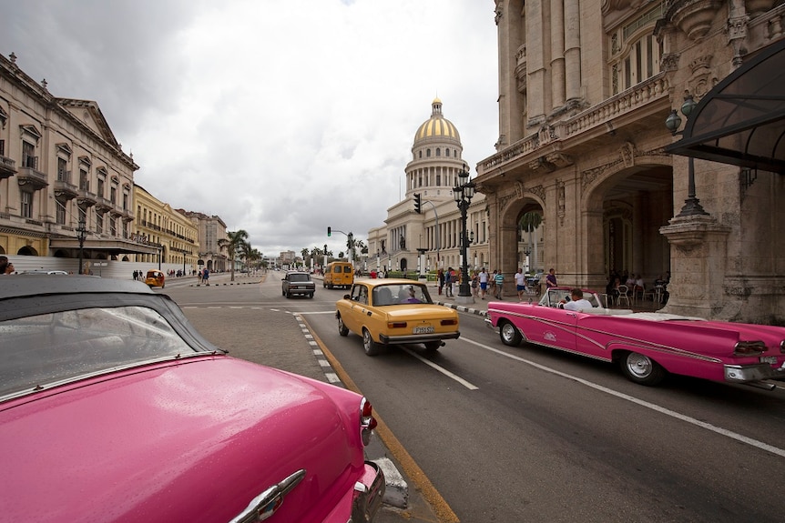 A bright pink vintage car and old taxi passes by the National Capitol Building in Havana on a rainy day.