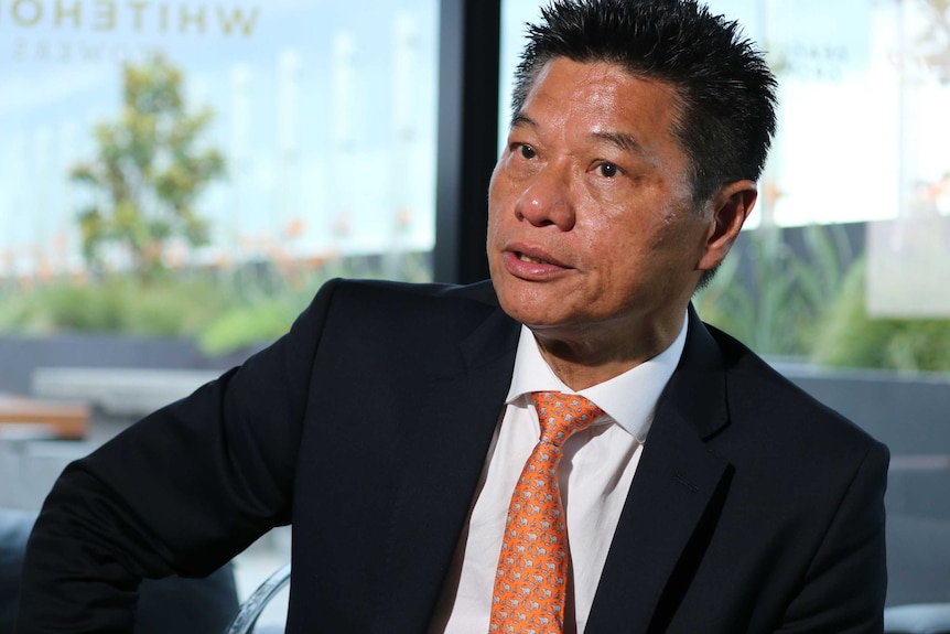 A man of Asian heritage with spiky black hair wears a black blazer and stares off camera.