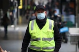 A man wearing a mask, a black cap and a fluro vest walks down the street.