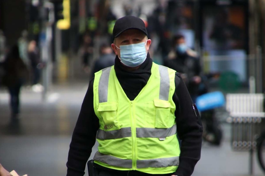 A man wearing a mask, a black cap and a fluro vest walks down the street.