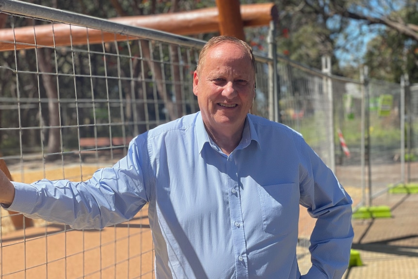 A man in a blue shirt stands in the sun holding on to a fence