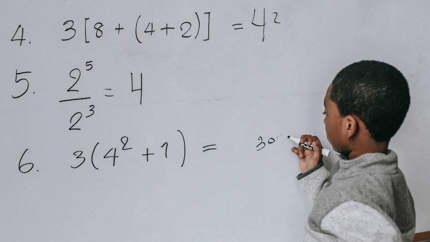 A boy holding a whiteboard pan and writing answers to maths equations on the board.