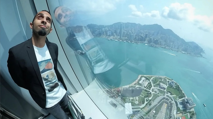 A man poses looking up as he stands in front of a window high above the city of Hong Kong in the background