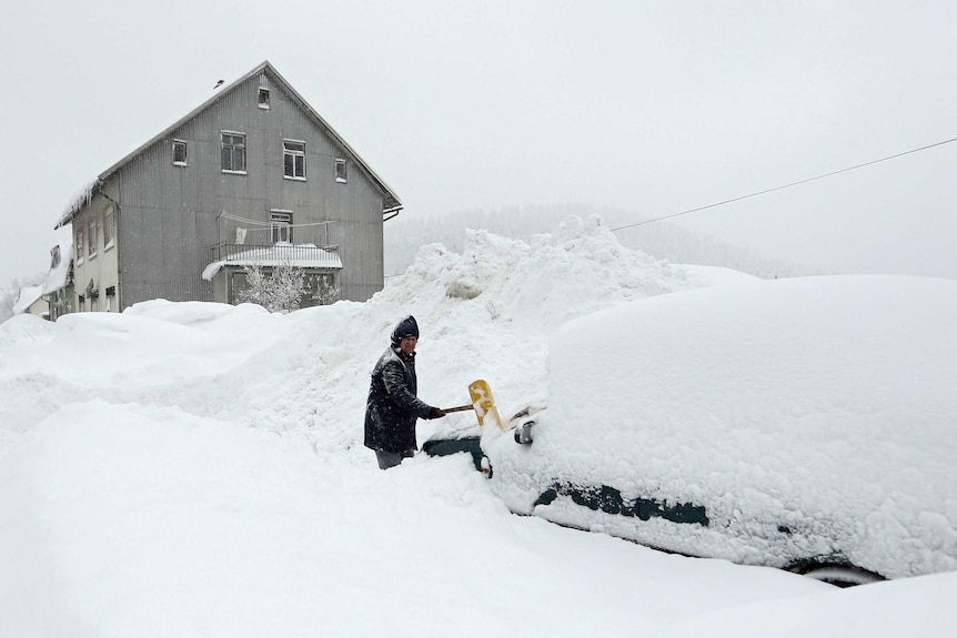 A man stands in thick snow while removing snow off his car.