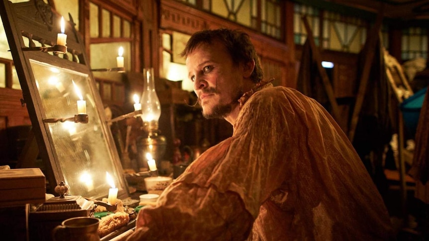 Damon Herriman, in costume, gazes into the distance while surrounded by candles