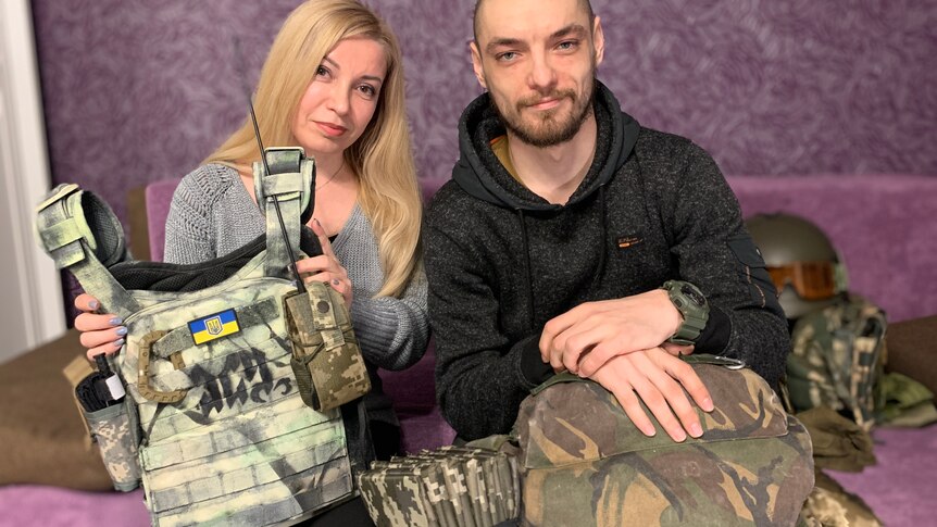 A woman and man hold up tactical military gear in a purple room.
