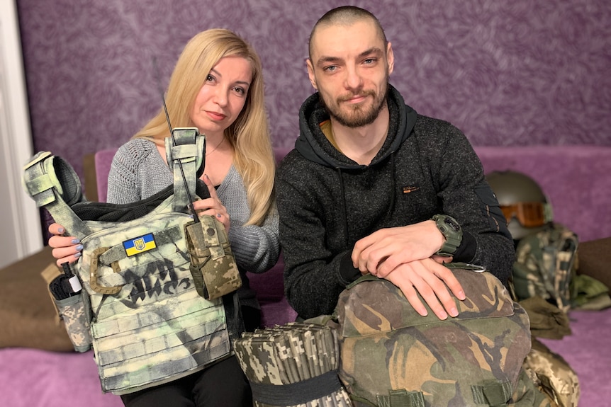 A woman and man hold up tactical military gear in a purple room.